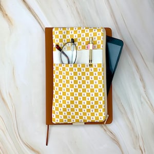 May include: A brown leather journal with a zippered pocket on the front. The pocket is lined with a yellow and white fabric with a small floral pattern. The journal is open and has a pen, glasses, and a tablet inside.