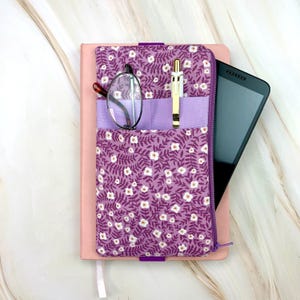 May include: A purple fabric cover with a white floral and leaf pattern, featuring a lavender pocket holding a pair of glasses and a gold pen. The cover is wrapped around a pink notebook and a black tablet, secured with a purple elastic band.