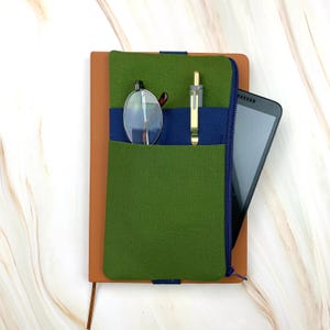 May include: A brown leather journal with a green felt pocket and a navy blue accent. The pocket holds a pair of eyeglasses, a gold pen, and a tablet. The journal is closed with a zipper.