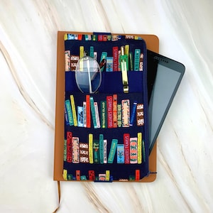 May include: A brown leather journal with a navy blue fabric cover featuring a colorful bookshelf pattern. The cover has pockets holding a pair of glasses, a gold pen, and a tablet. The journal is open, revealing the interior pages.