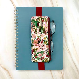 May include: A blue spiral-bound notebook with a red elastic band holding a floral print pen case and eyeglasses. The pen case features a zipper and holds three pens. The floral pattern includes pink and red roses.