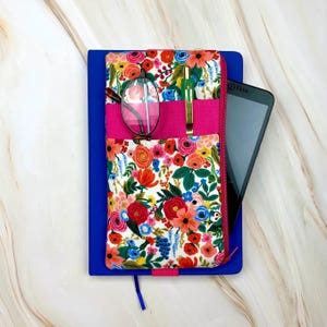 May include: A blue notebook with a floral print cover, a hot pink pocket, and a zipper. The pocket holds a pair of glasses, a gold pen, and a tablet. The floral pattern includes red, orange, and blue flowers.