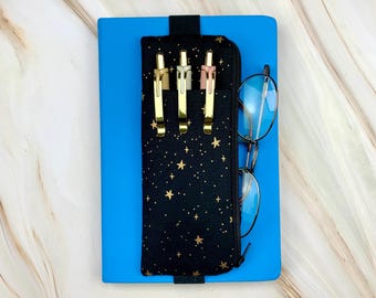 May include: A bright blue notebook with a black fabric pen case attached by an elastic band. The case features a gold star pattern and holds three pens with gold clips. A pair of thin-rimmed glasses is hooked onto the side of the pen case.