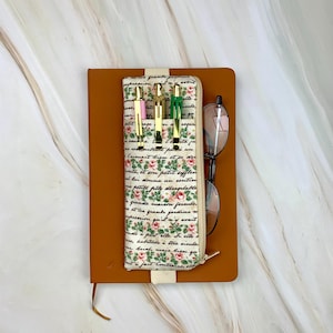May include: A brown leather journal with a floral print pen case containing three pens. The pen case has a cream background with pink and red roses and script. A pair of glasses rests on the journal.