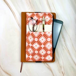 May include: A brown leather journal with a floral patterned pocket insert. The insert features a pink background with white daisy and brown leaf designs. The pocket holds a pair of glasses, a pen, and a smartphone.
