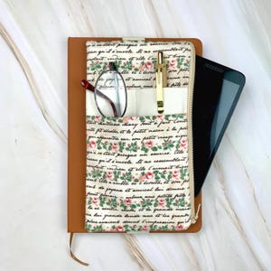 May include: A brown leather journal with a floral fabric pocket, holding eyeglasses, a gold pen, and a smartphone. The pocket features a cream-colored section and a floral pattern with text. A zipper secures the journal.