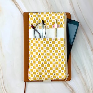 May include: A brown leather journal with a yellow and white checkered fabric cover. The cover has a pocket holding eyeglasses, a pen, and a smartphone. The fabric has a floral pattern. The journal is closed with a zipper.