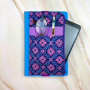 May include: A blue tablet case with a floral pattern in pink and blue. The case has a purple pocket holding glasses and a pen. A tablet is partially visible on the right side.