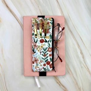 May include: A pink notebook with a black elastic band and a white ribbon bookmark. A floral print pen case with gold pens and a pair of eyeglasses rest on the notebook. The pen case has a white background with a colorful floral pattern.