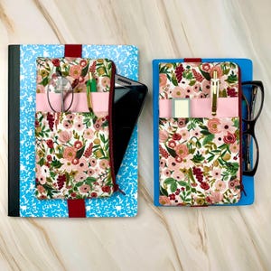 May include: Two notebooks with floral print covers. One notebook is blue with a white pattern, the other is blue. Each has a fabric pocket with a zipper, a pink band, and a red band. One has glasses, a pen, and a phone.