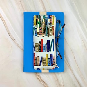 May include: A blue notebook with a cream-colored pencil case featuring a colorful book design. The pencil case holds several pens and a pair of eyeglasses. The zipper is gold-colored.