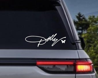 May include: A white vinyl decal applied to the rear window of a silver vehicle. The decal features the word Dolly written in a cursive script font, with a small butterfly icon positioned to the right of the text, creating a clean graphic design.