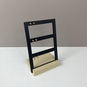 May include: A black earring display stand with a rectangular base and a vertical frame. The frame has multiple holes for hanging earrings. The base is a light beige colour. Gold earrings are displayed on the stand.