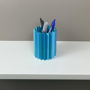 Può includere: Un portapenne blu turchese con un design geometrico a coste. Il contenitore cilindrico contiene una penna nera, una penna argentata e una penna blu. Il portapenne è su una superficie bianca con uno sfondo neutro.
