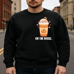 Op de afbeelding: Een zwart sweatshirt met ronde hals voorzien van een afbeelding van een oranje pompoen-ijskoffie in een doorzichtige plastic beker met een rietje. Het ontwerp bevat kleine roze hartjes en sterretjes, met de witte tekst ON THE ROCKS. eronder.