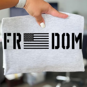 Op de afbeelding: Een lichtgrijs gemêleerd t-shirt met het woord FREEDOM in dikke, zwarte blokletters. De letter E is vervangen door een afbeelding van de Amerikaanse vlag, waarbij de sterren en strepen in zwarte inkt op de stof zijn gedrukt.