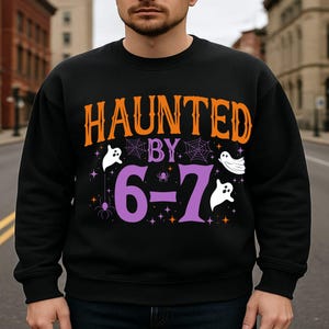 Peut inclure: Sweat-shirt noir avec l'inscription "HAUNTED BY 6-7" en orange et violet. Le motif comprend des fantômes blancs, des toiles d'araignées et des étoiles, idéal pour Halloween. Un haut décontracté pour la saison effrayante.