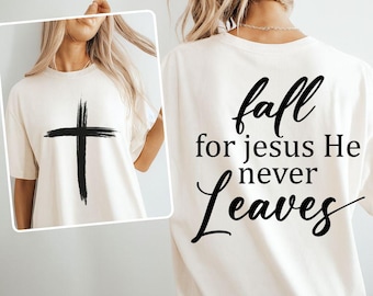 May include: Cream-colored t-shirt featuring a large black cross on the front. The back of the shirt displays the text "fall for Jesus He never Leaves" in a stylish, cursive font. A casual and comfortable design.
