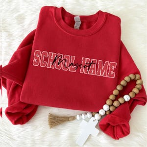 Op de afbeelding: Een felrood sweatshirt met ronde hals en de tekst SCHOOL NAME in een wit collegiaal lettertype met een zwarte cursieve overlay. Een snoer van houten kralen met een wit kruis en een beige kwastje ligt over de voorkant van het kledingstuk.