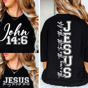 Op de afbeelding: Zwart T-shirt met witte tekst. De voorkant toont "John 14:6" in een scriptlettertype. De achterkant toont "JESUS the way the truth the life" verticaal in een lettertype in collegestijl. Een religieus kledingstuk.
