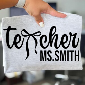 Op de afbeelding: Een lichtgrijs t-shirt met het woord "teacher" in zwart script, met een strik ontwerp. Daaronder staat de naam "MS. SMITH" in een vetgedrukte, zwarte letter. Het shirt wordt door een hand vastgehouden.