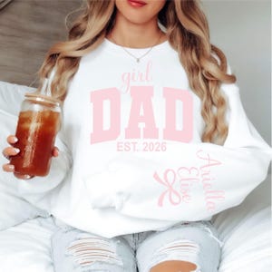 Op de afbeelding: Een wit sweatshirt met ronde hals en de lichtroze tekst girl DAD EST. 2026 op de borst. De linkermouw toont de namen Ariella en Elise in lichtroze cursief schrift, vergezeld van een kleine lichtroze strikgrafiek bij de boord.