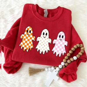 Op de afbeelding: Een rood sweatshirt met ronde hals en een afbeelding van drie geesten. De eerste geest heeft een oranje en wit dambordpatroon, de tweede is wit met kleine oranje pompoenen en de derde heeft een roze en wit dambordpatroon.