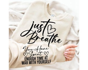 May include: A beige sweatshirt with the words "Just Breathe" and "You Have Spent Enough Time At War With Yourself" in black script. The sweatshirt is folded on a white surface.