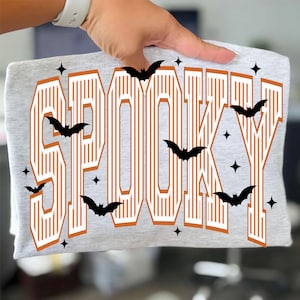 Op de afbeelding: Een lichtgrijs t-shirt met het woord SPOOKY in grote, oranje, varsity-stijl blokletters met witte verticale strepen. Het ontwerp is versierd met verschillende zwarte silhouetten van vliegende vleermuizen en kleine vierpuntige sterren.