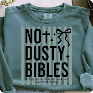 Op de afbeelding: Een tealkleurige sweatshirt met de tekst "NOT DUSTY BIBLES" in zwart, versierd met een strik en sterren. Het ontwerp bevat ook de zin "No Dusty Bible, No Empty Faith, Just A Heart Filled With God's Word" eronder.