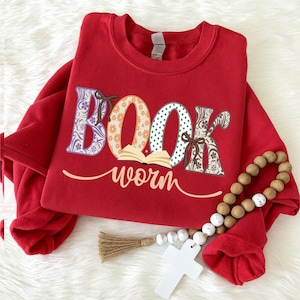 Op de afbeelding: Een rood sweatshirt met ronde hals voorzien van het woord BOOK in grote, gedessineerde letters, met een kleine afbeelding van een open boek eronder en het woord worm in cursief schrift. Een houten kralenslinger met een wit kruis ligt erop.