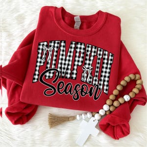 Op de afbeelding: Een rood sweatshirt met ronde hals voorzien van de tekst PUMPKIN in een zwart-wit ruitpatroon met kleine witte strikjes, en het woord Season in een zwart cursief lettertype. Het sweatshirt wordt getoond met een houten kralenslinger.