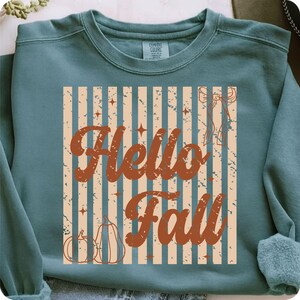 Op de afbeelding: Een blauwgroene sweater met ronde hals en een grafische print met verticale beige strepen en een verweerde textuur. Het ontwerp bevat de bruine tekst Hello Fall, twee omlijnde pompoenen, kleine vierpuntige sterren en een strik.