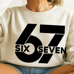 Puede incluir: Sudadera color crema con el número "67" en negrita negra. La palabra "SIX" está colocada debajo del "6" y "SEVEN" debajo del "7". La sudadera es de estilo casual y cómodo.