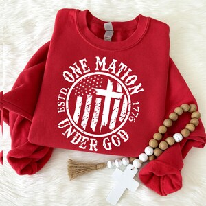 Op de afbeelding: Een rood sweatshirt met ronde hals en een witte cirkelvormige afbeelding met de tekst "ESTD. ONE NATION UNDER GOD 1776" rondom een gestileerde Amerikaanse vlag en een kruis. Een snoer van houten kralen met een wit kruis hangt over de stof.