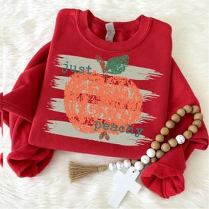 Op de afbeelding: Een rode sweatshirt met de tekst "just peachy" en een verweerde perzikafbeelding. Het ontwerp bevat grijze horizontale strepen achter de perzik. Een houten kralenkrans en een wit kruis staan op de voorgrond.