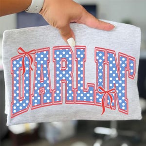 Op de afbeelding: Een grijs t-shirt met het woord "DARLIN" in grote, vette letters. De letters zijn blauw met witte stippen en rood omlijnd. Rode strikken zijn aan weerszijden van de tekst geplaatst.