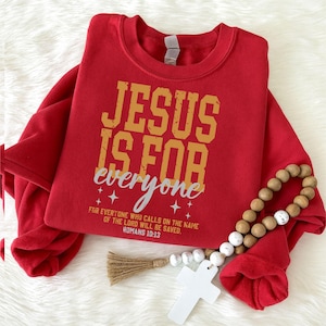 Op de afbeelding: Een rood sweatshirt met ronde hals voorzien van de gele blokletters JESUS IS FOR en de witte cursieve tekst everyone. Daaronder staat de tekst FOR EVERYONE WHO CALLS ON THE NAME OF THE LORD WILL BE SAVED. ROMANS 10:13 met kleine ster-iconen.