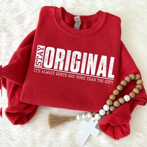 May include: A bright red crewneck sweatshirt featuring white distressed-style text that reads STAY ORIGINAL IT'S ALWAYS WORTH WAY MORE THAN THE COPY. The garment includes a long-sleeved design with a ribbed collar and a decorative wooden bead strand.