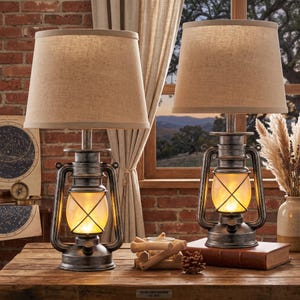 May include: Two table lamps featuring a rustic, weathered silver-tone metal lantern base with a glowing amber light inside. Each lamp is topped with a tapered, light beige fabric drum shade. The lamps sit on a wooden surface with a pinecone.