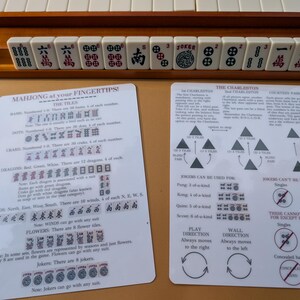 Pode incluir: Um conjunto de Mahjong com pe&ccedil;as brancas com s&iacute;mbolos pretos e vermelhos, dispostas em um suporte de madeira. Duas folhas de instru&ccedil;&otilde;es detalham as regras e estrat&eacute;gias do jogo. O texto "MAHJONG at your FINGERTIPS!" &eacute; vis&iacute;vel.