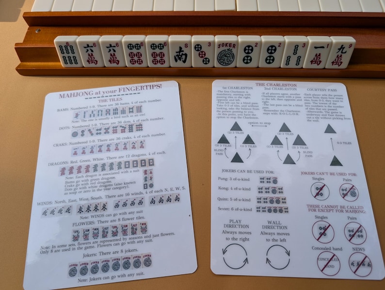 Pode incluir: Um conjunto de Mahjong com pe&ccedil;as brancas com s&iacute;mbolos pretos e vermelhos, dispostas em um suporte de madeira. Duas folhas de instru&ccedil;&otilde;es detalham as regras e estrat&eacute;gias do jogo. O texto "MAHJONG at your FINGERTIPS!" &eacute; vis&iacute;vel.