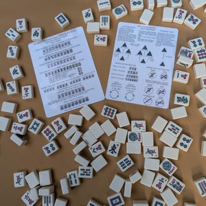 Pode incluir: Um conjunto de jogo de Mahjong com pe&ccedil;as de cor marfim espalhadas sobre uma superf&iacute;cie bege. As pe&ccedil;as apresentam v&aacute;rios s&iacute;mbolos e caracteres em vermelho, verde e preto. Duas folhas de instru&ccedil;&otilde;es tamb&eacute;m s&atilde;o vis&iacute;veis, com texto e diagramas. O conjunto &eacute; um jogo de tabuleiro cl&aacute;ssico.