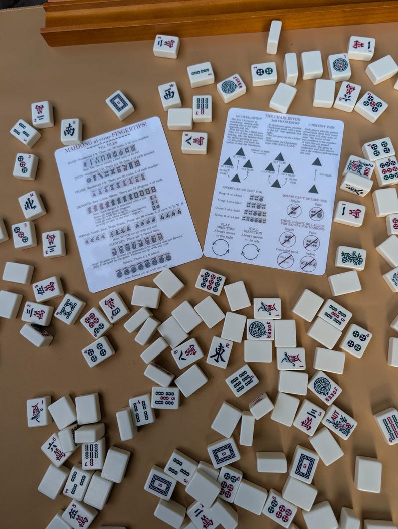 Pode incluir: Um conjunto de jogo de Mahjong com pe&ccedil;as de cor marfim espalhadas sobre uma superf&iacute;cie bege. As pe&ccedil;as apresentam v&aacute;rios s&iacute;mbolos e caracteres em vermelho, verde e preto. Duas folhas de instru&ccedil;&otilde;es tamb&eacute;m s&atilde;o vis&iacute;veis, com texto e diagramas. O conjunto &eacute; um jogo de tabuleiro cl&aacute;ssico.