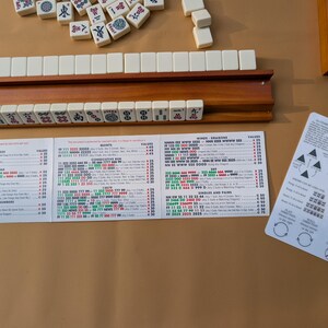 Pode incluir: Um conjunto de Mahjong com pe&ccedil;as de cor marfim, um suporte de madeira e cart&otilde;es de regras impressos. As pe&ccedil;as est&atilde;o dispostas sobre uma superf&iacute;cie castanha. Os cart&otilde;es de regras apresentam instru&ccedil;&otilde;es e diagramas detalhados.
