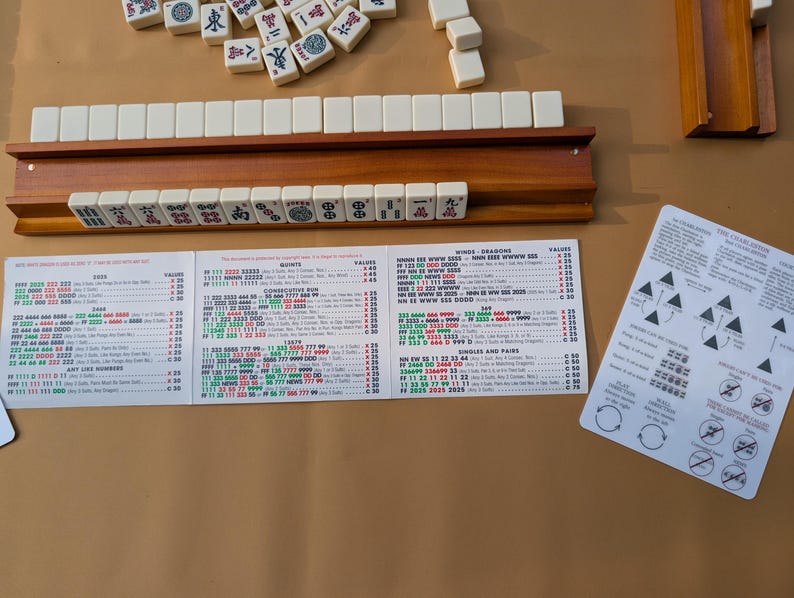 Pode incluir: Um conjunto de Mahjong com pe&ccedil;as de cor marfim, um suporte de madeira e cart&otilde;es de regras impressos. As pe&ccedil;as est&atilde;o dispostas sobre uma superf&iacute;cie castanha. Os cart&otilde;es de regras apresentam instru&ccedil;&otilde;es e diagramas detalhados.