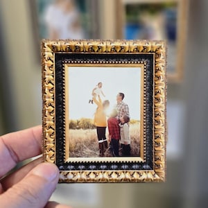 May include: A small, ornate gold picture frame with a black border, containing a family portrait. The frame is held in a hand, with two other framed pictures in the background. The family in the photo is standing in a field.