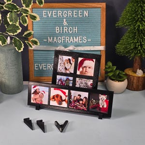 Puede incluir: Dos marcos de fotos negros con múltiples aberturas, cada uno con fotos pequeñas. Los marcos se exhiben sobre una superficie azul claro, con un letrero de fondo que dice "Evergreen & Birch Magframes". Elementos decorativos incluyen un pequeño árbol de Navidad y una suculenta.
