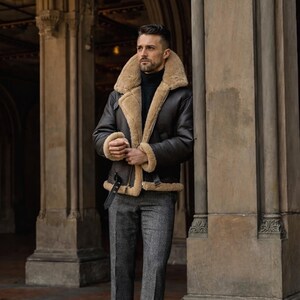 Pu&ograve; includere: Giacca in montone marrone scuro con fodera e colletto in montone beige. La giacca ha una cintura nera e viene indossata con pantaloni di tweed grigi. Le misure della giacca non sono visibili.