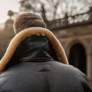 Pu&ograve; includere: Una giacca di pelle nera con colletto in montone beige vista da dietro. La giacca ha una cerniera e una finitura strutturata. I capelli della persona sono visibili e lo sfondo &egrave; sfocato, suggerendo un ambiente esterno.
